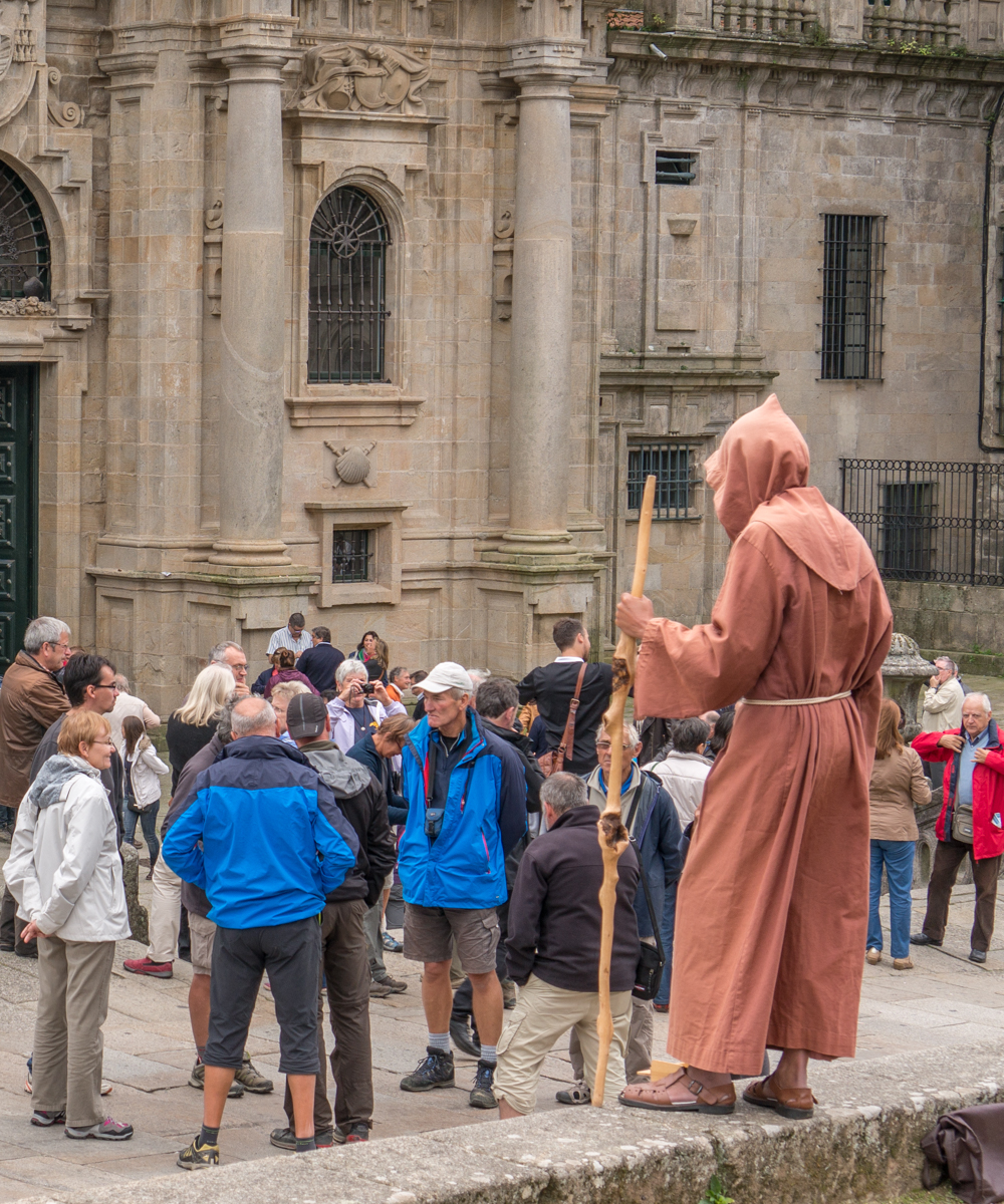 Contemporary Camino pilgrims exiting the Catedral de Santiago (Spain) at completion of the noon Mass walk past a local resident dressed as a typical medieval pilgrim | Photo by Mike Hudak
